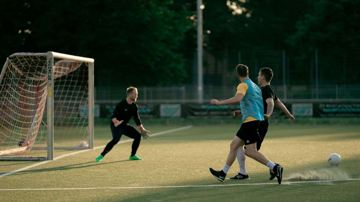 Two soccer players and a goalie in action during an outdoor match at sunset.