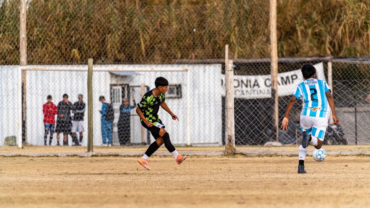 Two teenagers playing soccer on an outdoor field with onlookers behind a fence.