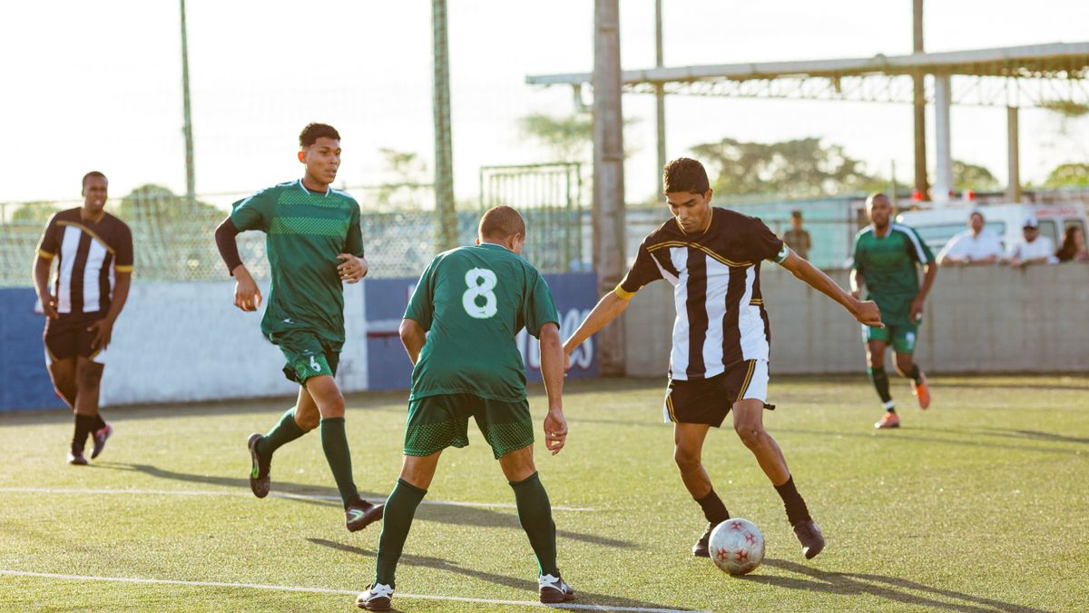 Five male soccer players in action during a daytime soccer match on an outdoor field.