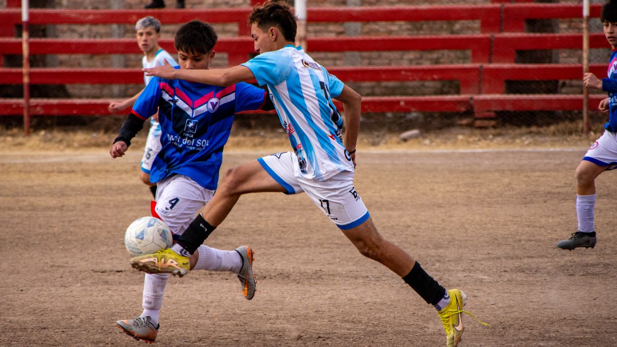 Teen soccer players compete fiercely during a match on a dirt field with red bleachers.