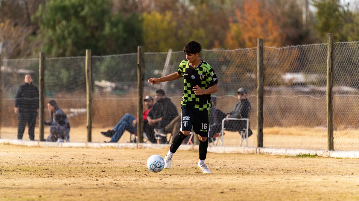 A young soccer player dribbles on a field, surrounded by spectators, on a sunny day.