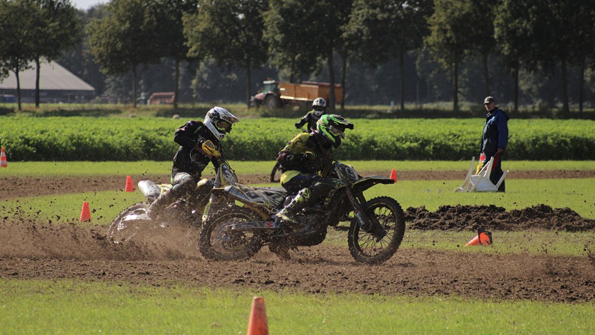 Two motorcyclists racing on a grass track with spectators and cones in the background.