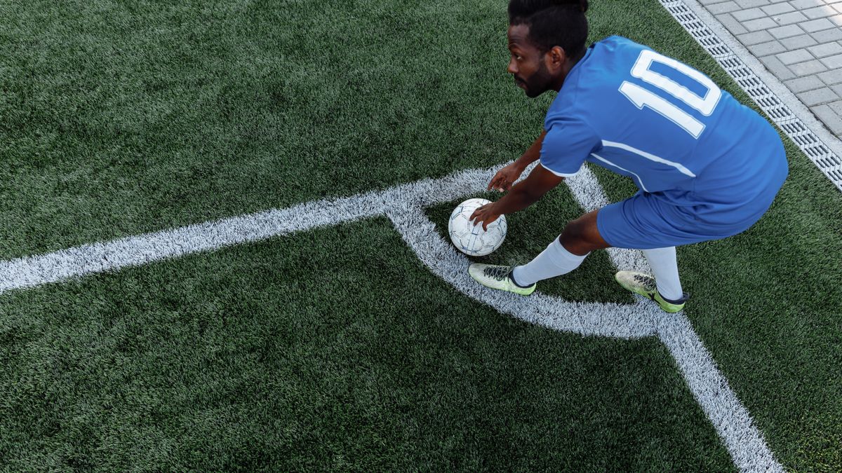 A soccer player in a blue jersey prepares to kick the ball from the corner on an artificial turf field. A soccer player in a blue jersey prepares to kick the ball from the corner on an artificial turf field.