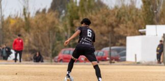 A soccer player wearing number 6 dribbling during a daytime match on a dry, grassy field.
