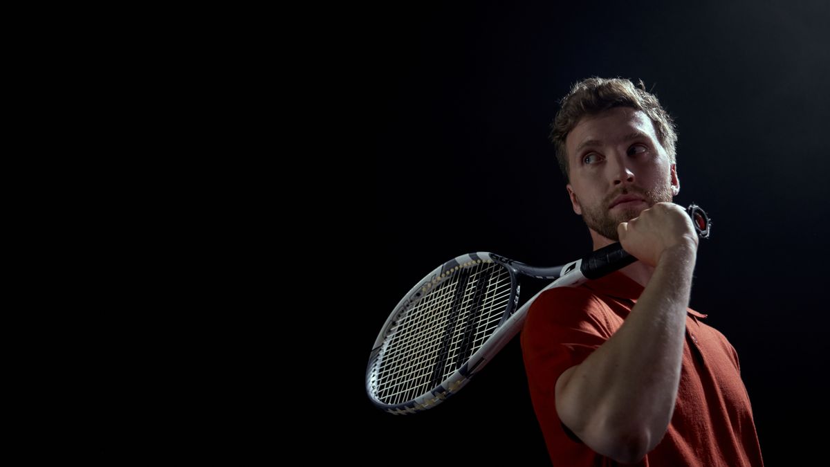 A thoughtful male tennis player holding a racket, set against a dark background. A thoughtful male tennis player holding a racket, set against a dark background.