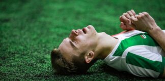Young male soccer player in green jersey lying on grass, eyes closed, exhausted.