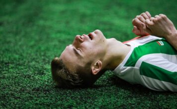 Young male soccer player in green jersey lying on grass, eyes closed, exhausted.