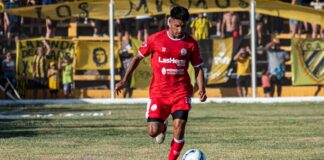A soccer player in red uniform dribbling during a daytime match with spectators.