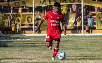 A soccer player in red uniform dribbling during a daytime match with spectators.
