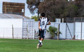 A teenage soccer player wearing number six dribbles the ball outdoors in a park.