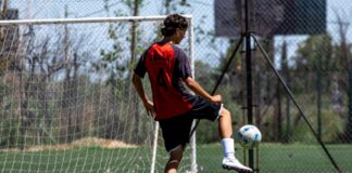 Teenager in red jersey playing soccer on an outdoor field during daytime.