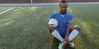 African man in blue jersey sitting on grass field holding a soccer ball.