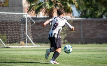 A soccer player in action, wearing a striped jersey, kicking a ball towards the goal during a sunny day match.