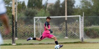 A young goalkeeper in pink uniform kicks a soccer ball on an outdoor field.