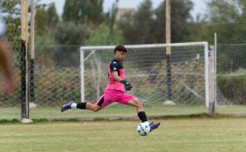A young goalkeeper in pink uniform kicks a soccer ball on an outdoor field.