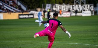 A goalkeeper in bright pink kit kicking a soccer ball during a match.