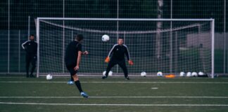 Male football player kicking ball towards goalkeeper standing against goal during training in field