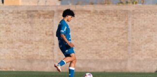A teenage soccer player in blue kit practicing on a green field by a brick wall.