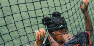 Back view of African American teenage football player sitting on field ground and holding grid of gate while preparing to start play football match