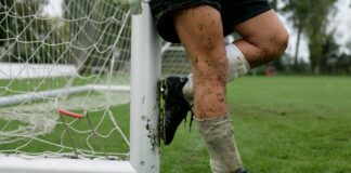 A soccer player with muddy legs leaning against a goal post on a grassy field.
