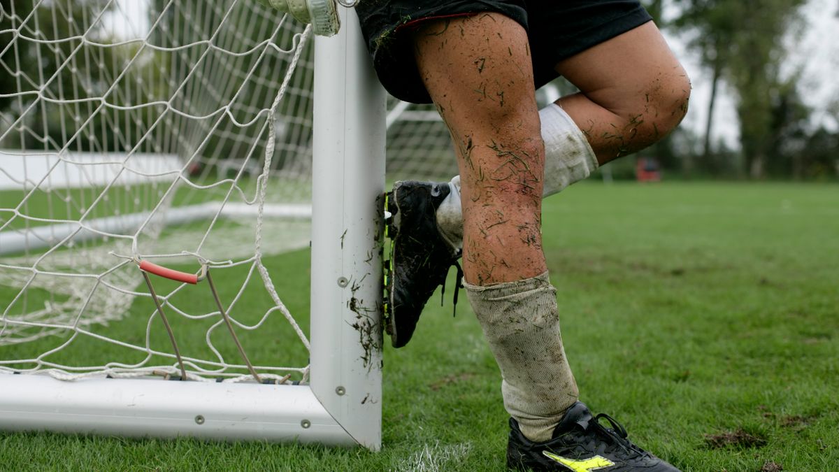 A soccer player with muddy legs leaning against a goal post on a grassy field. A soccer player with muddy legs leaning against a goal post on a grassy field.