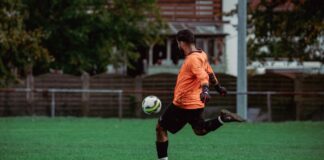 Soccer player in orange jersey kicking a ball on a lush green field during a daytime game.