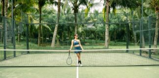 A woman poses with a tennis racket on a palm tree-lined tennis court outdoors.