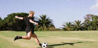 A woman soccer player kicking a ball on a grassy field during a sunny day outdoors.