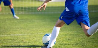 A young soccer player in blue uniform kicks a ball towards the goal on a sunny day.