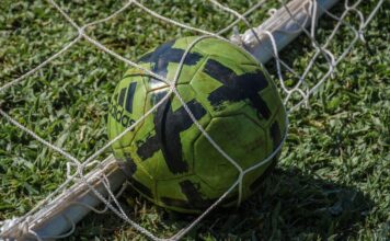 Bright green soccer ball stuck in a net on a grassy field in sunlight.