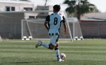 A young soccer player wearing a number 8 jersey runs with the ball on a sunny outdoor field.