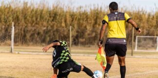 Soccer player tackles during a match as referee observes. Captured outdoors on a field.
