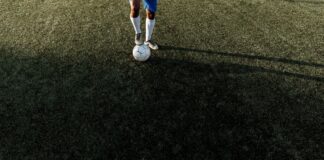 A soccer player stands on a field with a ball under bright sunlight, casting a long shadow.