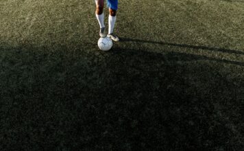 A soccer player stands on a field with a ball under bright sunlight, casting a long shadow.