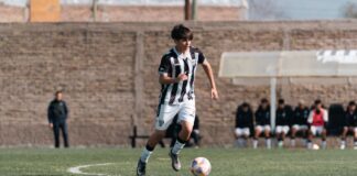 Teenage soccer player dribbling on a sunny day during a match with teammates in the background.