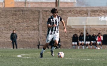 Teenage soccer player dribbling on a sunny day during a match with teammates in the background.