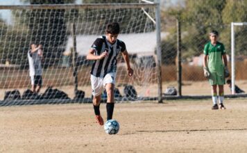 Teen soccer player dribbling the ball during a match on a sunny day outdoors.