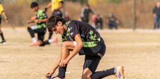Teenage soccer player kneeling, adjusting cleats during a match on a sunny day.