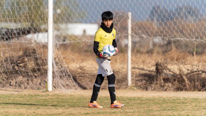 A young soccer goalkeeper standing on the field, holding a ball during the day. A young soccer goalkeeper standing on the field, holding a ball during the day.